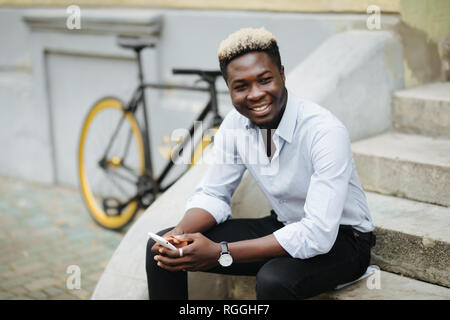 Bello Afro American uomo in abbigliamento casual si utilizza lo smartphone mentre è seduto sulle scale all'aperto Foto Stock