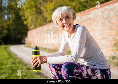 Sorridente sportive donna senior tenendo la bottiglia in posizione di parcheggio Foto Stock