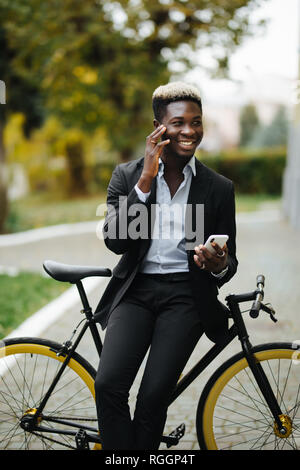 Bel giovane africano indossare tuta di parlare al telefono mentre si cammina con la bicicletta al lavoro la mattina. Imprenditore di andare a lavorare con la sua bicicletta Foto Stock