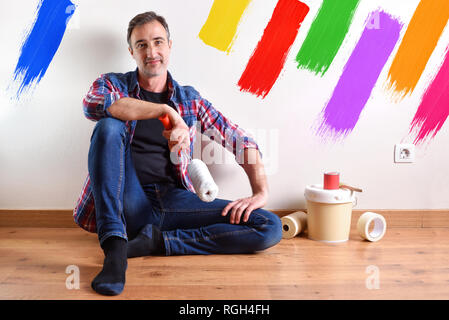 Uomo seduto su un pavimento in parquet preparati con materiale di vernice per ridipingere la sua casa e la parete dipinta con sette colori. Vista frontale. Compositio orizzontale Foto Stock