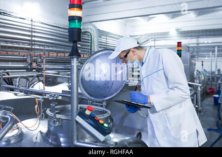 Il Tecnologo in camice bianco e la maschera rende note con la penna in foglio in corrispondenza del caseificio. Vista di profilo caseificio fabbrica Lavoratore che indossa nel complesso permanente al processo di produ Foto Stock