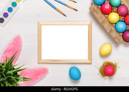 Un telaio, orecchie di coniglietto, un vaso di fiori artificiali con erba verde e colorati di uova in una scatola di cartone, vernici e pennelli. Concetto di pasqua. Spazio di copia Foto Stock