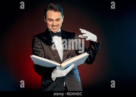 Professional mago tuta da indossare guanti di nd in piedi isolato su nero e sfondo rosso azienda libro magico incantesimo rendendo sorridente eccitato Foto Stock