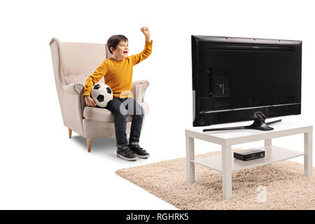 A piena lunghezza shot di un ragazzino seduto in poltrona con pallone da calcio e guardare una partita in tv isolato su sfondo bianco Foto Stock