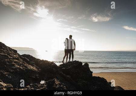 Francia, Bretagna, vista posteriore della coppia giovane permanente sulla roccia in spiaggia al tramonto Foto Stock