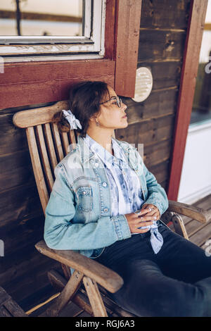 La Svezia, Lapponia, pensieroso giovane donna seduta su una sedia sulla veranda che guarda a distanza Foto Stock
