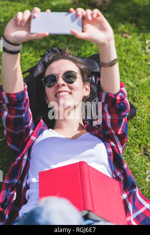 Vista dall'alto di un giovane hipster sorridente Donna sdraiata sull'erba in una giornata di sole in un parco mentre si prende un selfie con un telefono cellulare Foto Stock