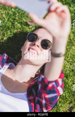 Vista dall'alto di un giovane hipster sorridente Donna sdraiata sull'erba in una giornata di sole in un parco mentre si prende un selfie con un telefono cellulare Foto Stock