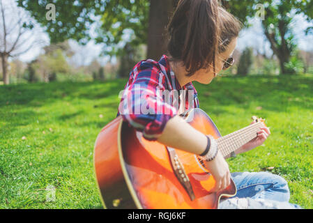 Vista frontale di una giovane donna hippster che indossa occhiali da sole, seduta sull'erba in un parco mentre si gode la chitarra Foto Stock