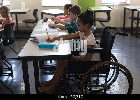 Vista laterale della scolaretta disabili con i compagni di classe di studiare e seduti alla scrivania in aula Foto Stock