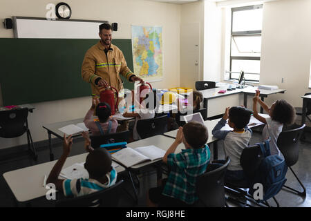 Vista frontale di un schoolkids applaudire mentre caucasico maschio pompiere insegnamento circa la sicurezza antincendio in aula della scuola elementare Foto Stock