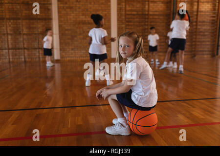 Vista laterale di schoolgirl seduta sul basket e guardando la telecamera a pallacanestro Foto Stock