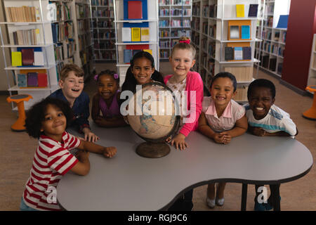 Vista frontale di felice schoolkids con globo guardando la fotocamera in biblioteca scolastica Foto Stock