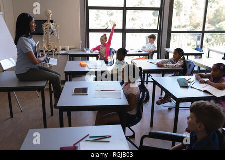 Vista laterale della scuola femminile insegnante schoolkids insegnamento sul computer portatile in aula della scuola elementare Foto Stock