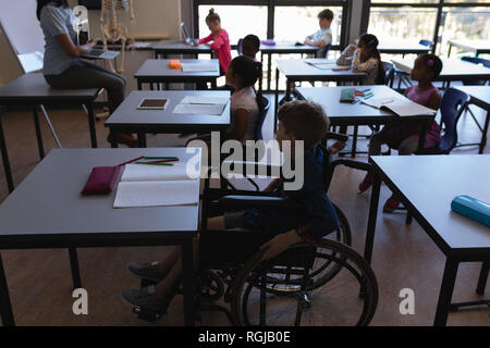 Vista laterale di disabilitare scolaro con i compagni di classe di studiare e seduti alla scrivania in classe della scuola elementare Foto Stock