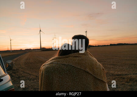 Giovane avvolto in una coperta a camper van nel paesaggio rurale con le turbine eoliche in background Foto Stock