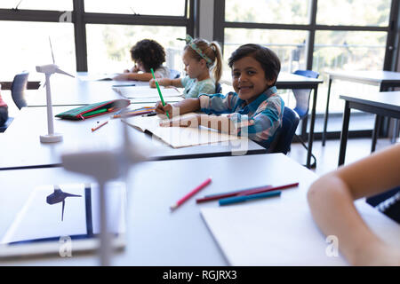 Vista laterale di scolaro seduto alla scrivania e guardando la telecamera in aula della scuola elementare Foto Stock