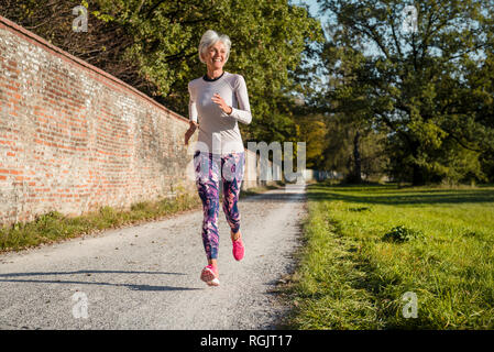 Senior donna che corre lungo un muro di mattoni in un parco Foto Stock