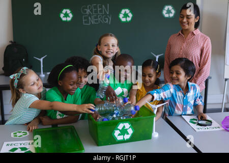 Vista frontale del schoolkids mettendo le bottiglie nel contenitore di riciclo al banco in aula della scuola elementare Foto Stock