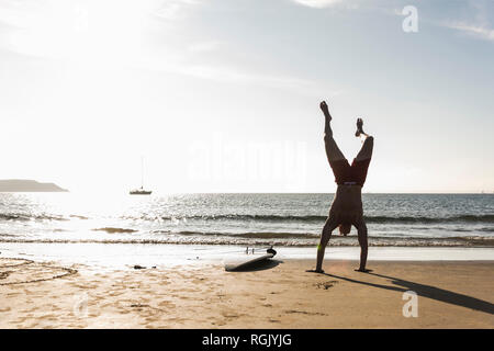 Francia, Bretagna, giovane uomo facendo un handstand sulla spiaggia accanto alla tavola da surf Foto Stock