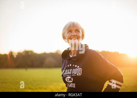 Senior sorridente donna che indossa una felpa con cappuccio in piedi sul prato rurale al tramonto Foto Stock