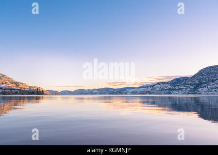 Cielo sereno con i colori del tramonto riflesso nelle calme acque del lago, con vista di lontane montagne coperte di neve Foto Stock