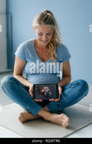 Sorridente donna incinta seduta sul pavimento che mostra l'immagine ad ultrasuoni su tablet Foto Stock