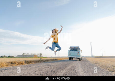 Esuberante giovane donna saltando su sterrato a camper van nel paesaggio rurale Foto Stock
