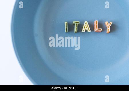 La parola "l'Italia" composta con pasta reale lettere in bandiera italiana i colori in un piatto blu. Sfondo con copia spazio sulla sinistra. Foto Stock