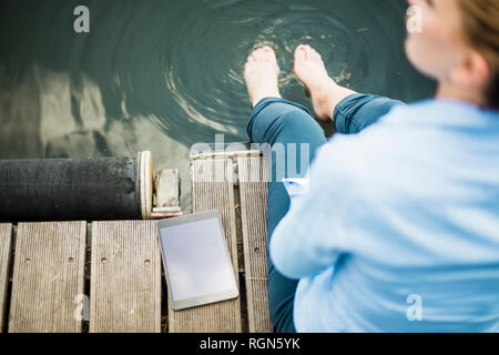 Donna seduta sul pontile di un lago vicino al tablet con i piedi in acqua Foto Stock