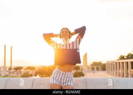 Spagna, Barcellona, Montjuic felice giovane donna l'ascolto di musica con le cuffie al tramonto Foto Stock