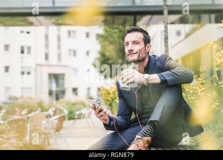 Imprenditore seduto fuori, prendendo una pausa, ascoltando la musica dal suo smartphone, bere caffè Foto Stock