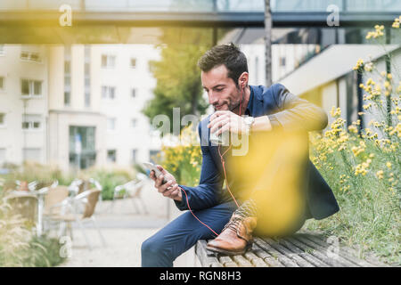 Imprenditore seduto fuori, prendendo una pausa, ascoltando la musica dal suo smartphone, bere caffè Foto Stock