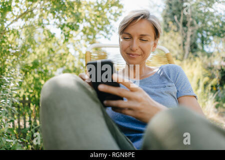 Donna seduta in giardino su una sedia con il telefono cellulare Foto Stock