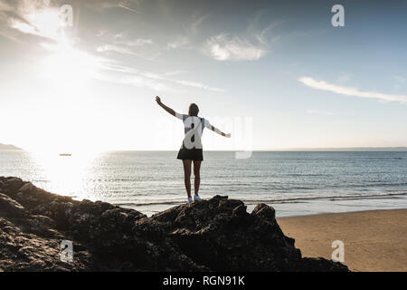 Francia, Bretagna, vista posteriore della giovane donna in piedi sulla roccia in spiaggia al tramonto Foto Stock