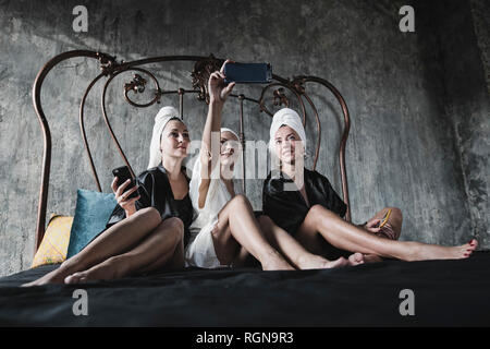 Tre donne con asciugamani intorno ai suoi capi sul letto prendendo un selfie Foto Stock