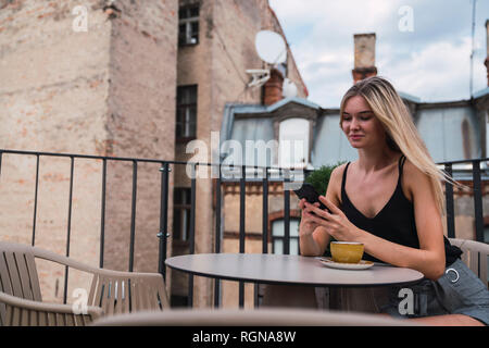 Sorridente donna bionda seduti sulla terrazza sul tetto con la tazza di caffè controllo telefono cellulare Foto Stock