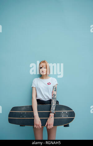 Ritratto di giovane donna holding carver skateboard in piedi alla parete turchese Foto Stock