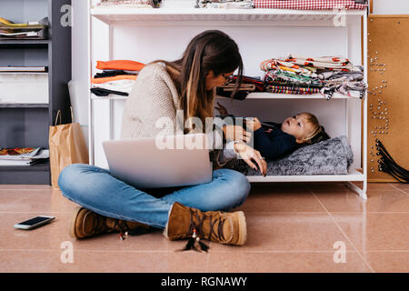 Madre seduta sul pavimento di casa utilizzando laptop con piccola figlia giacente nel ripiano Foto Stock