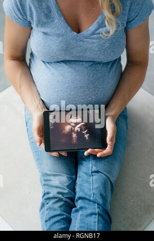 Donna incinta seduta sul pavimento che mostra l'immagine ad ultrasuoni su tablet Foto Stock