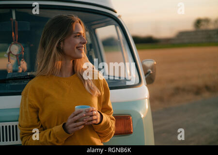 Felice giovane donna azienda tazza in camper nel paesaggio rurale al tramonto Foto Stock