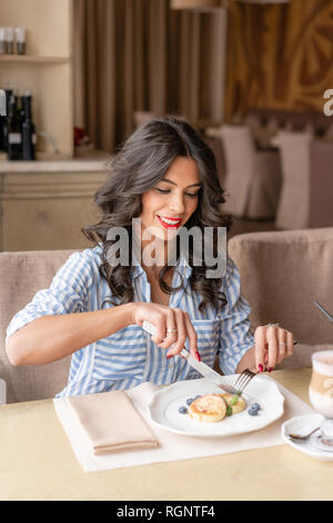 Bella giovane donna tagli frittelle con la lama di un coltello, godendo di caffè cappuccino o latte in un bicchiere di vetro con schiuma. Colazione Leggera vicino alla finestra in un cafe Foto Stock
