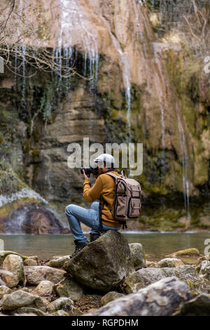 Vista posteriore del viaggiatore maschio con seduta sulle pietre e scattare foto con la fotocamera del fantastico cascata nella foresta in Spagna Foto Stock