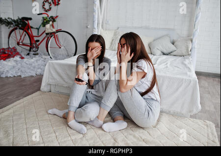 Due gemelli modelle Ragazze mentre fai clic sul telecomando del TV su studio e nascondere i volti, perchè hanno guardato il film spaventoso. Sono spaventato un Foto Stock