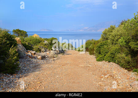 Strada di campagna di pietre e sabbia, sui lati dei cespugli verdi, in lontananza sul mare e sulle montagne Foto Stock