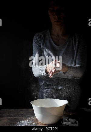 Crop lady battendo le mani nella farina vicino a tazza, confezione di uova e frusta sul tavolo di legno su sfondo nero Foto Stock