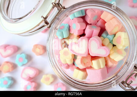Una coppia di cuore rosa sulla pila di pastello a forma di fiore Marshmallow caramelle in un vaso di vetro con alcuni sparsi sul tavolo e il concetto di San Valentino Foto Stock