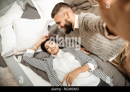 Attraente dai capelli scuri rilassante donna sul letto mentre il marito facendo foto Foto Stock