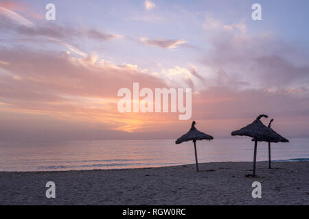 Inverno pallido tramonto di mare luci e soffice clounds con delicati colori rosa, arance e i colori dei coralli. Tre con tetto di paglia di ombrelloni da spiaggia in primo piano. Foto Stock