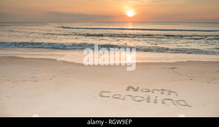Le parole Carolina del Nord inciso nella sabbia al sunrise con onde che arrivano a riva. Foto Stock
